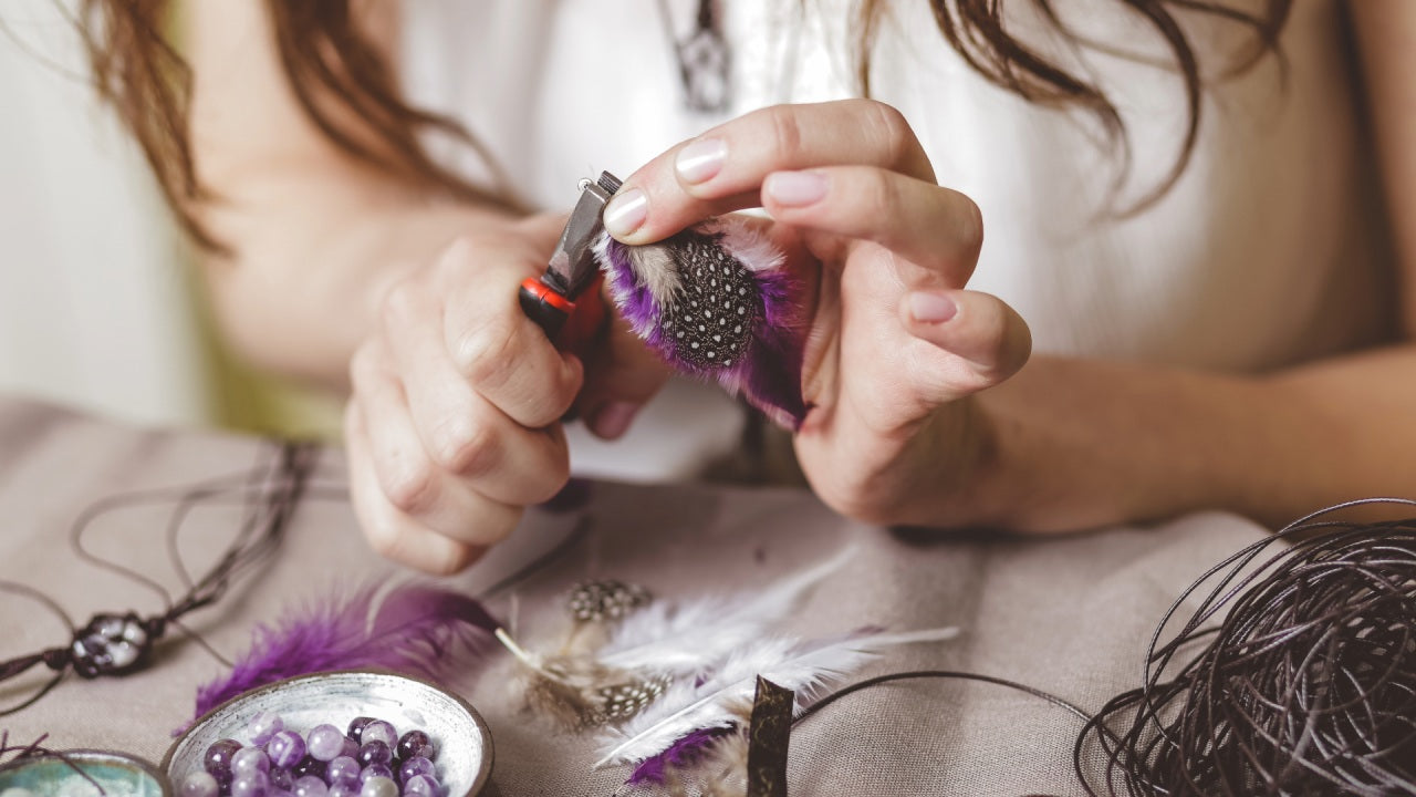 Crafter using jewelry pliers to assemble handmade accessories with purple dyed and dotted feathers for craft projects at a workspace with beads and cords.